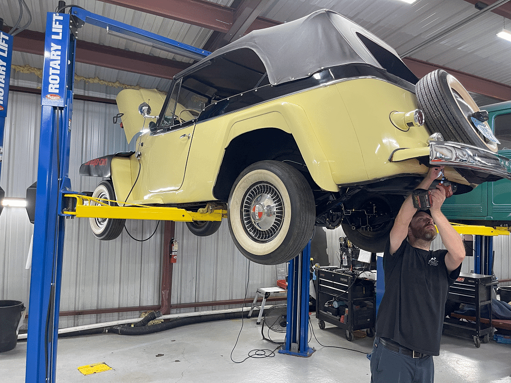 Classic 1948 Willys Jeepster Overland being serviced in a restoration shop, lifted on a Rotary Lift, with a technician using a power tool for maintenance.